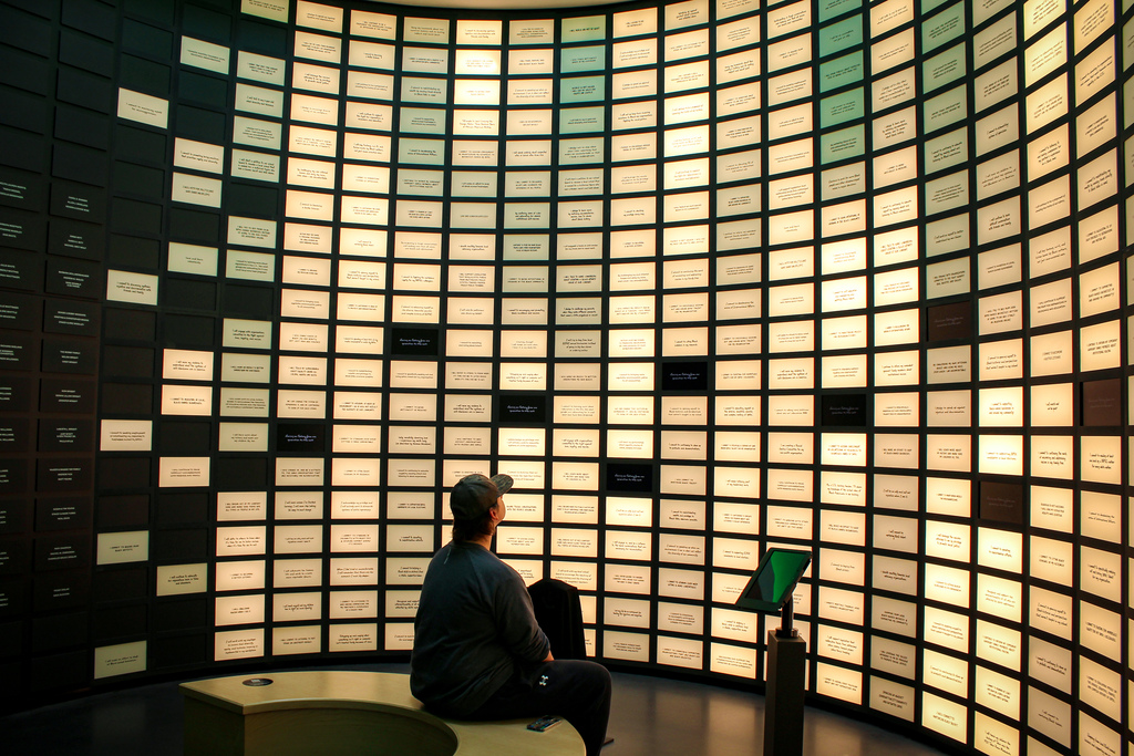 Person in a museum or exhibit, seated, facing a curved wall of light panels and a small monitor.