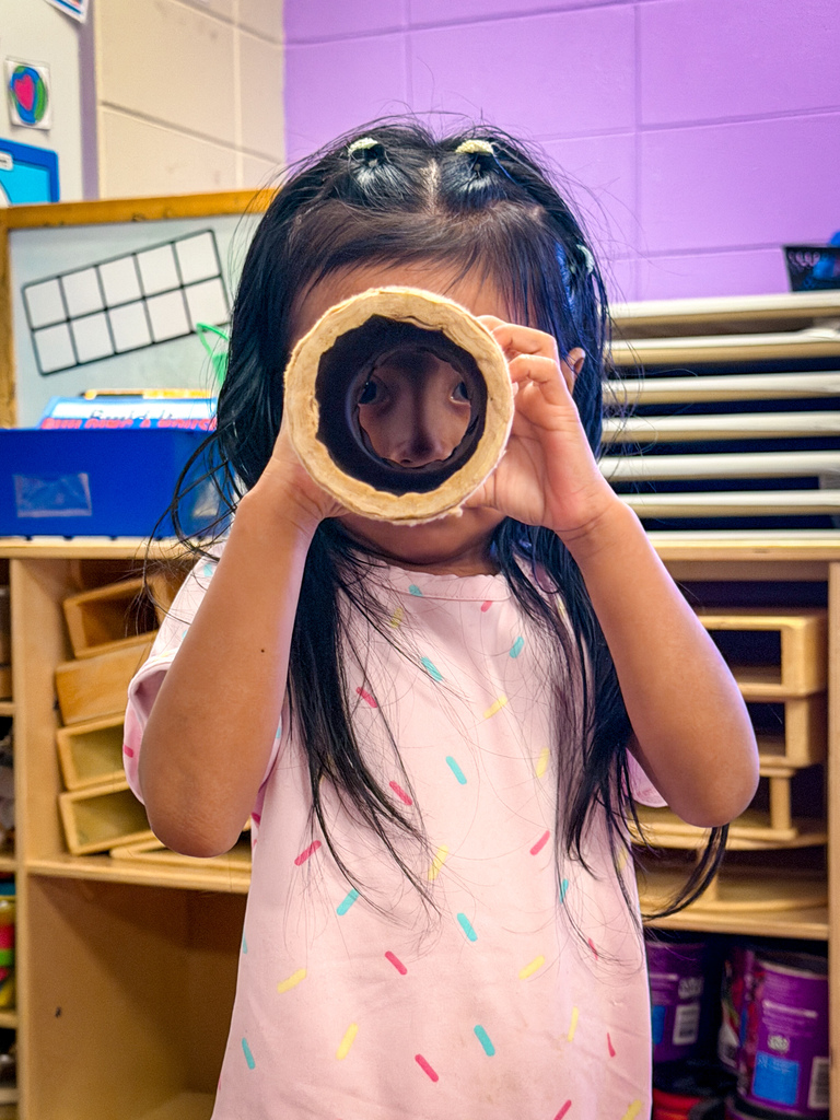 A young girl with long hair stands in a classroom, holding a rolled-up paper tube up to her face.