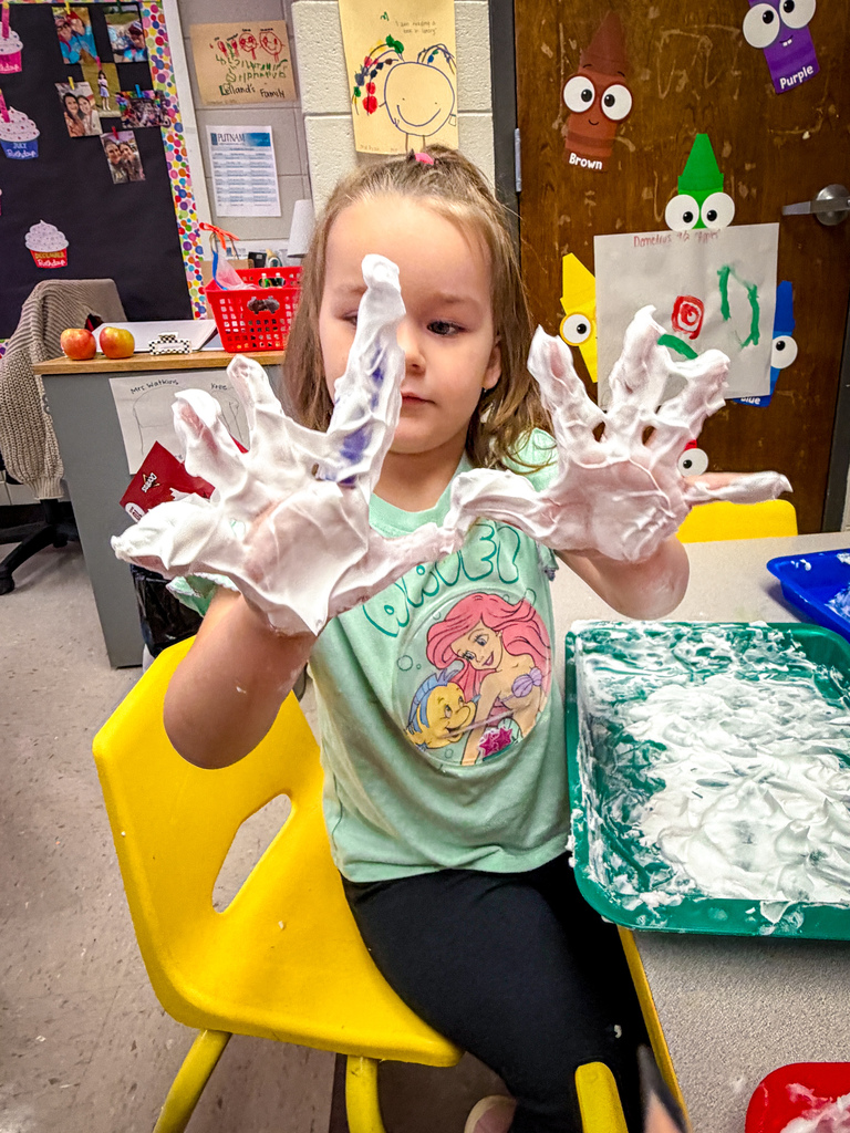 A young girl with blue hair sits at a table with foamy hands, in a classroom.