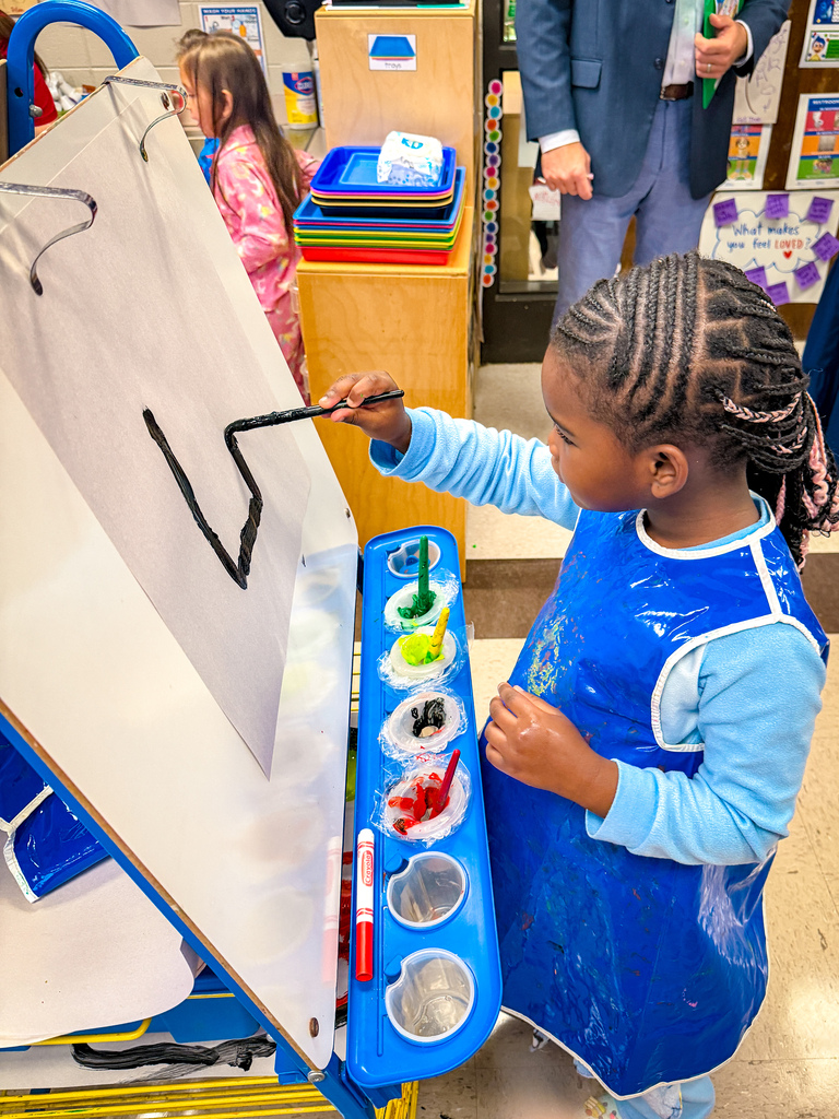 Child painting an upside down V on a large vertical paper board with paintbrush.