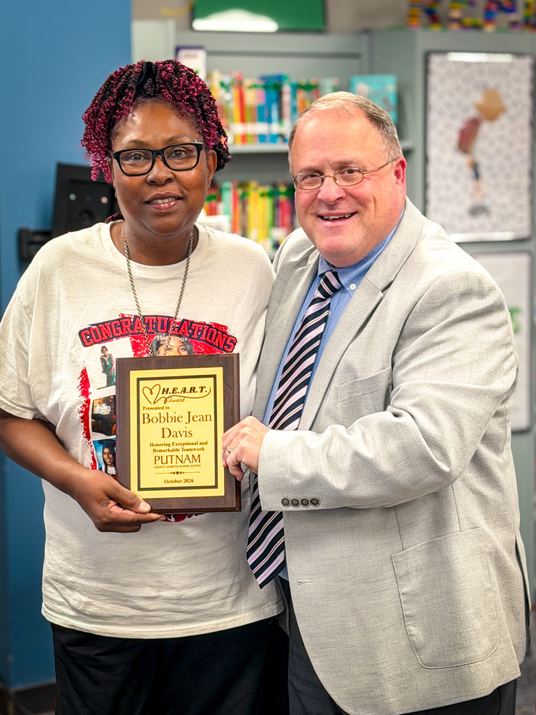 A woman and a man stand in a library with a blue background. The woman holds a plaque, and the man smiles.