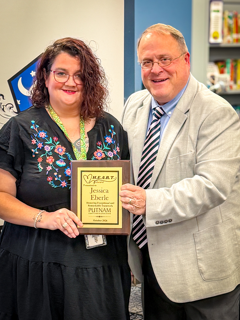 A woman in a floral shirt holds a plaque while a man in a suit smiles. They stand in an indoor area with shelves and a wall design in the background.