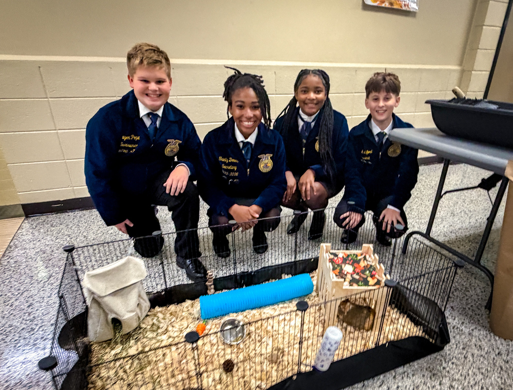 Five children in matching uniforms crouch in front of a guinea pig habitat, with a small house, water bottle, and tunnel visible.