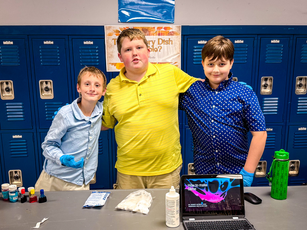 Three boys in light blue, yellow, and dark blue shirts stand behind a table in a school hallway.
