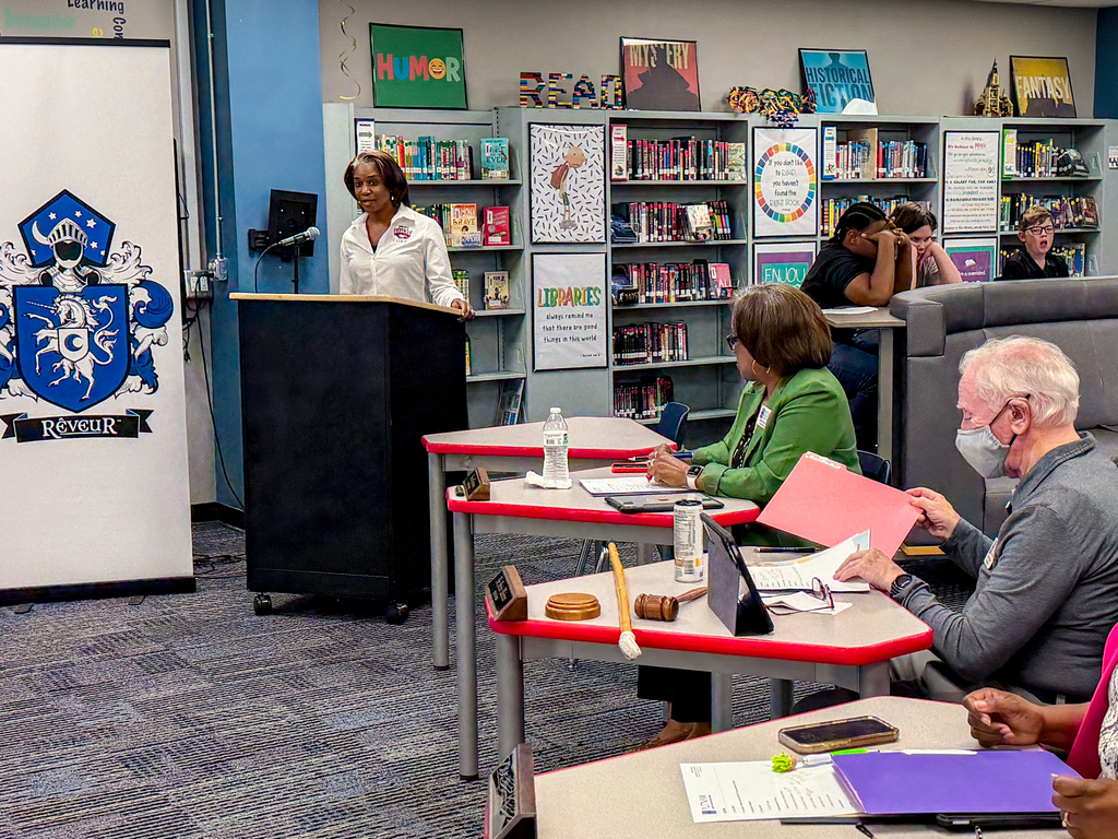 Several people sit at tables, one with a podium and mask, in a library with books and a blue wall.