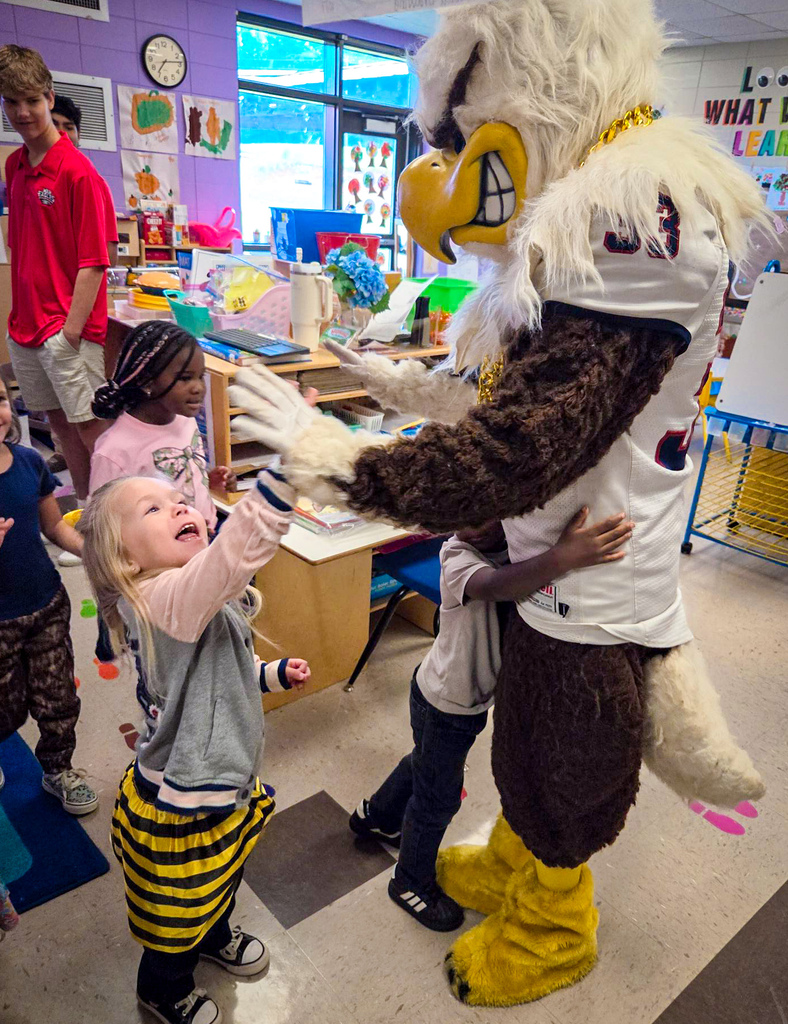 A mascot resembling an eagle wearing a white jersey with black numbers engages with children in a classroom.