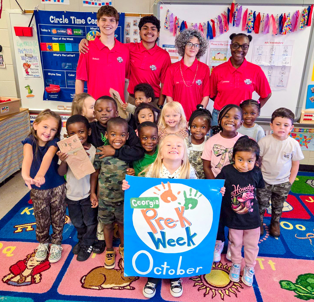 Preschool students and staff in red shirts, holding signs, posing for a photo in a classroom.