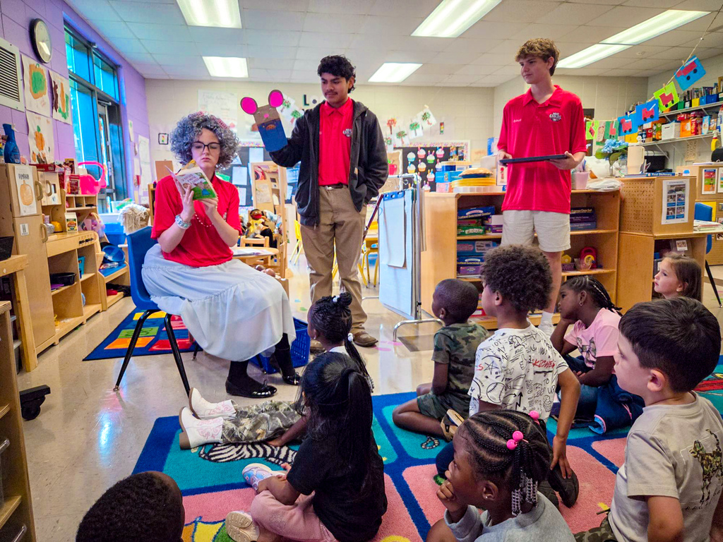 Children seated on the floor, attentively listening to a woman in a chair, with two men in red shirts standing.