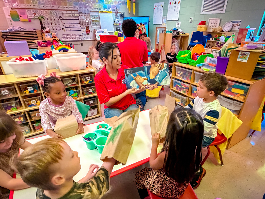 A group of children sit at a table in a classroom. One child holds a book and a paper bag.