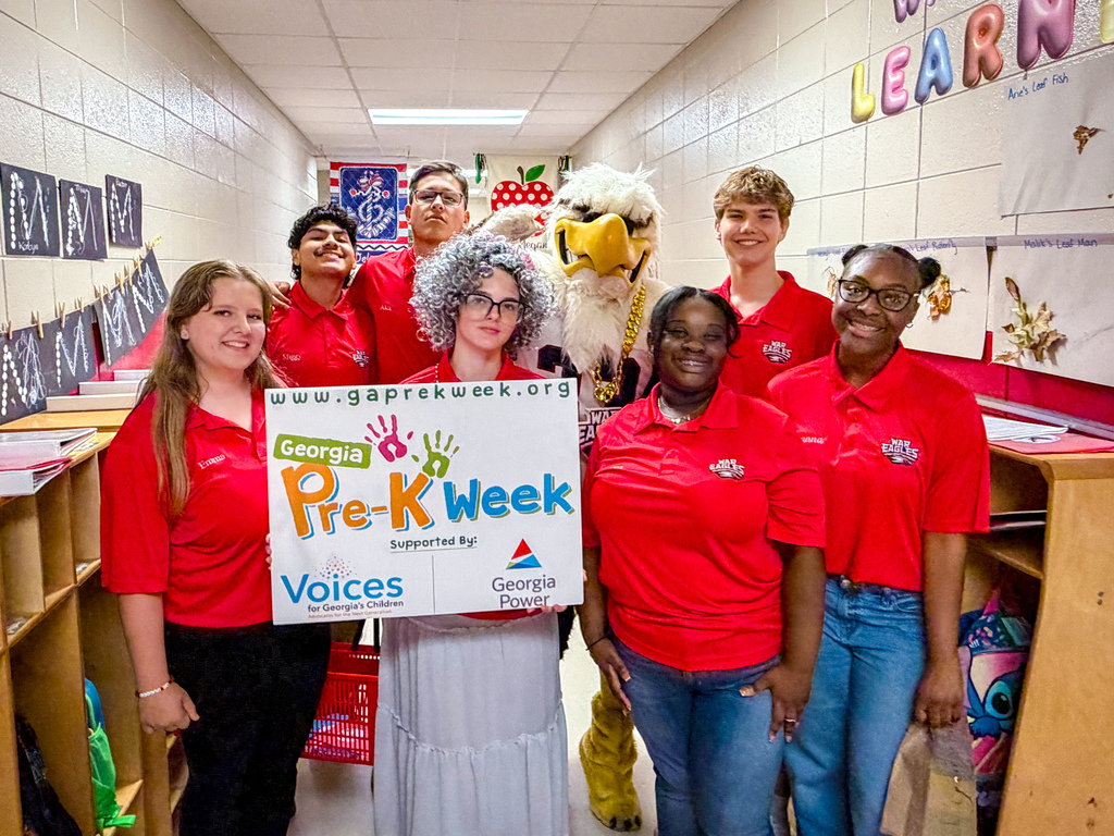 Group of adults in red shirts stand in a hallway holding a sign that says "Pre-K Week."