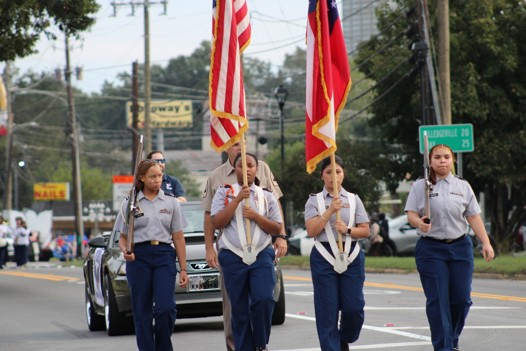 Four women in matching uniforms march in a parade, carrying flags. A car is parked behind them.