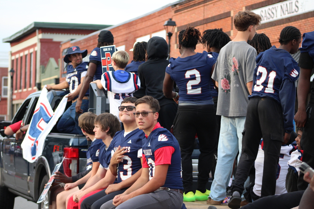 Group of people in sports uniforms. Several on a pickup truck, others standing. Bright, sunny day.