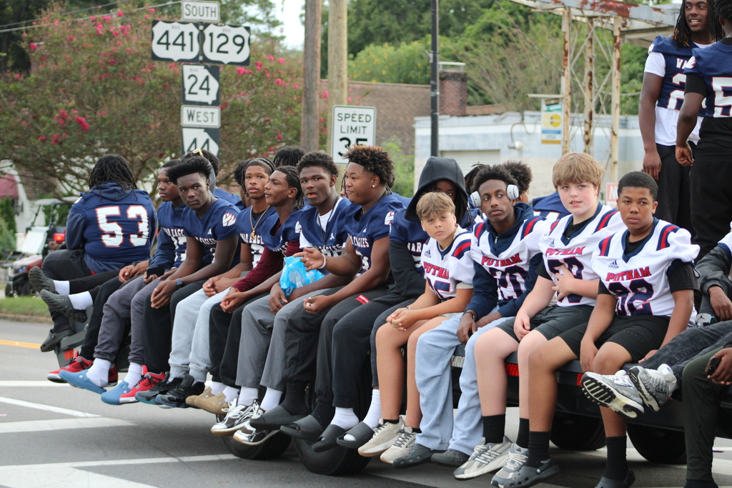 A group of football players in blue and white uniforms sit on the back of a truck.