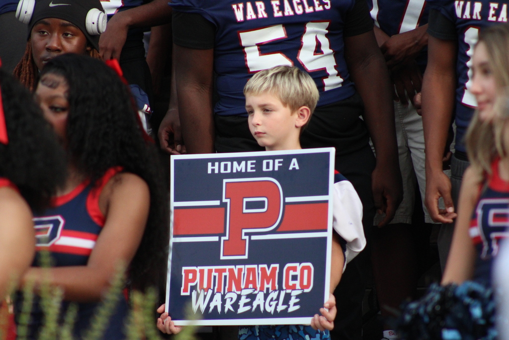 A boy holds a sign reading "Home of a P" in front of cheering people in sports gear.