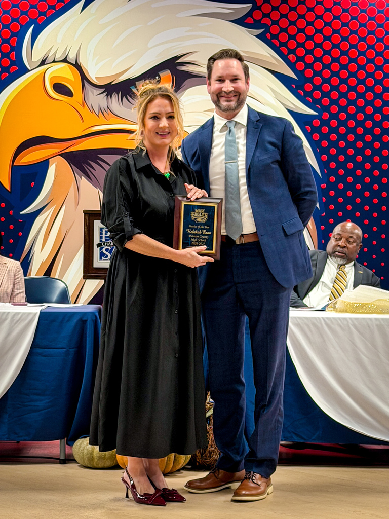 Woman in black dress holding plaque with man in blue suit and tie in front of red, white, and blue backdrop.