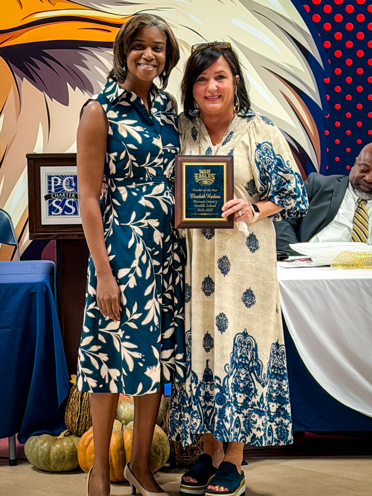 Two women stand in front of a wall with an eagle design, holding an award plaque. A man in a suit sits behind a table.