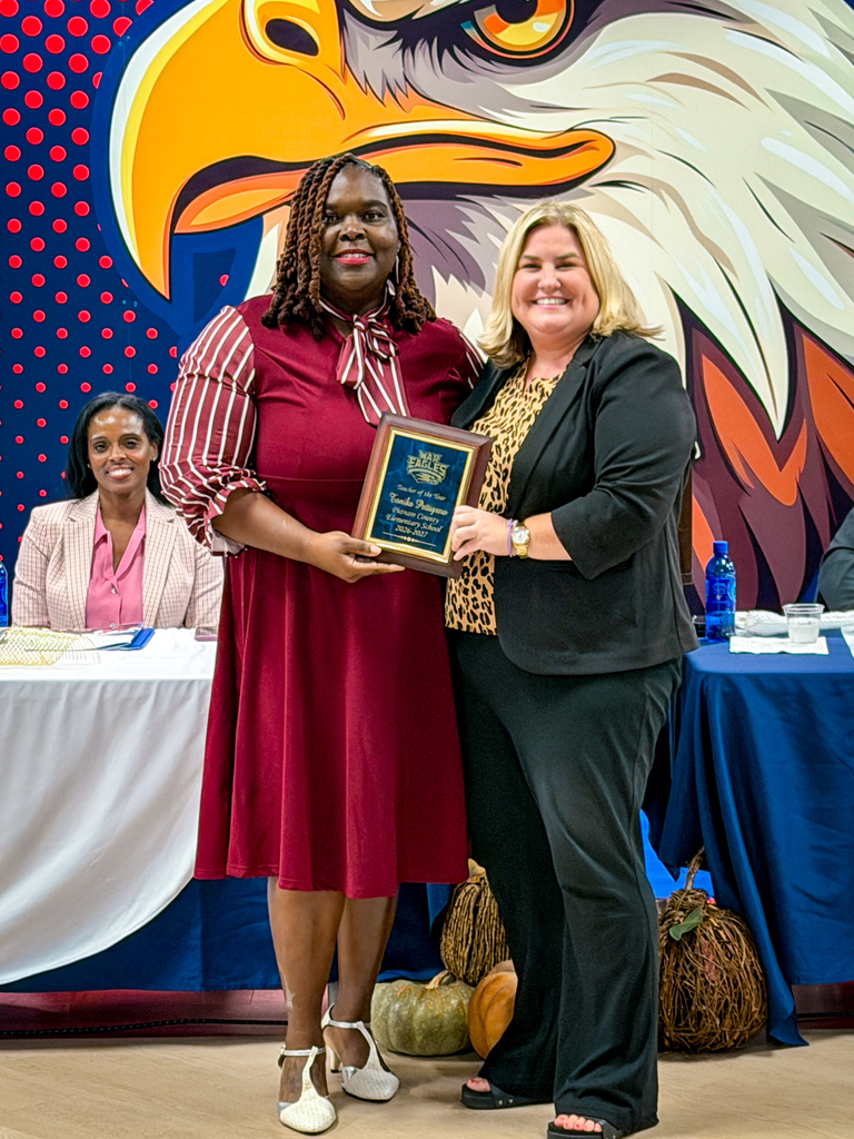 Two women standing, one holding framed plaque, with large eagle mural and table with pumpkins behind them.