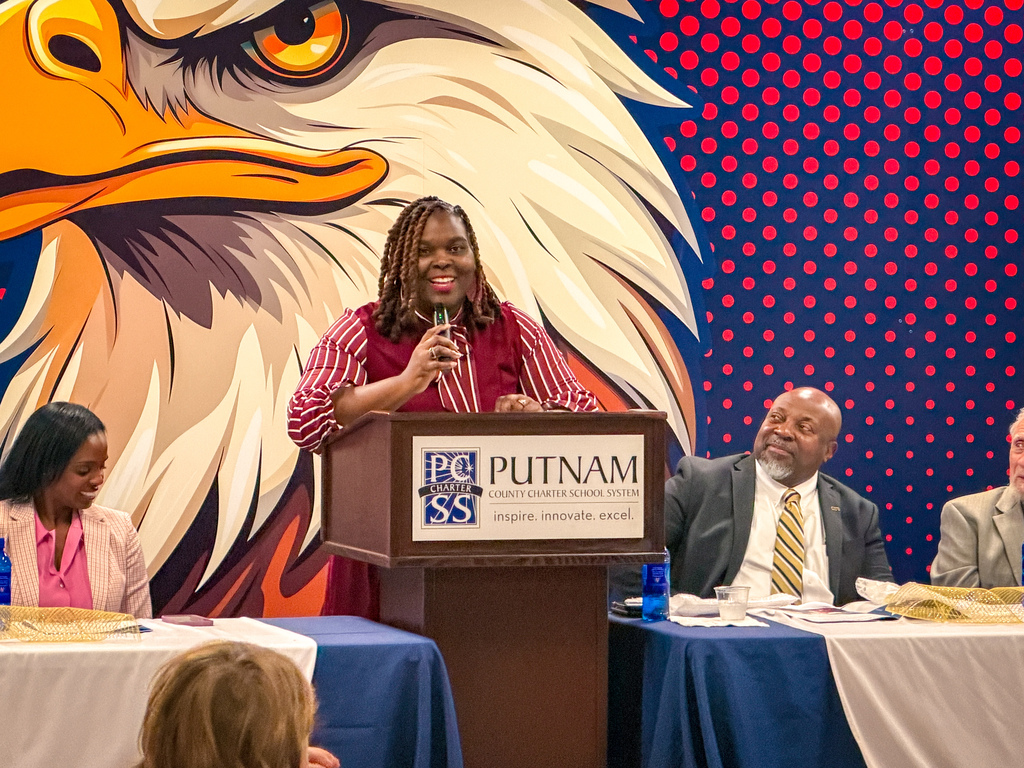 Woman at podium with microphone, three people seated behind her. Eagle on wall behind podium.