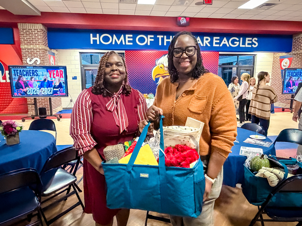 Two women hold a blue bag filled with items, one with glasses. Tables with blue tablecloths and chairs behind them.