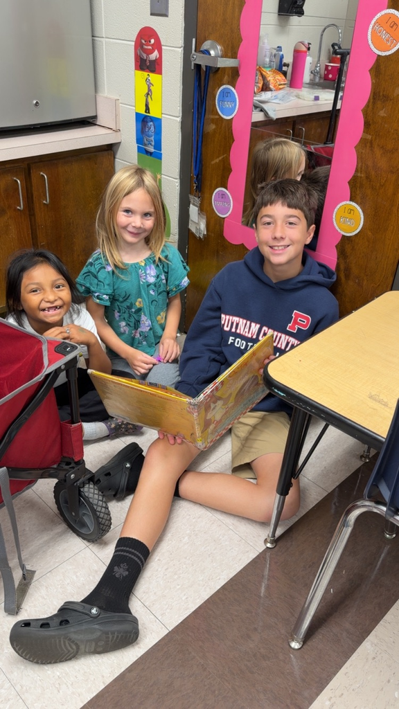 Three children sit in a classroom; two are reading a book while a boy smiles. Behind them are cabinets, a refrigerator, and a mirror with a reflection.