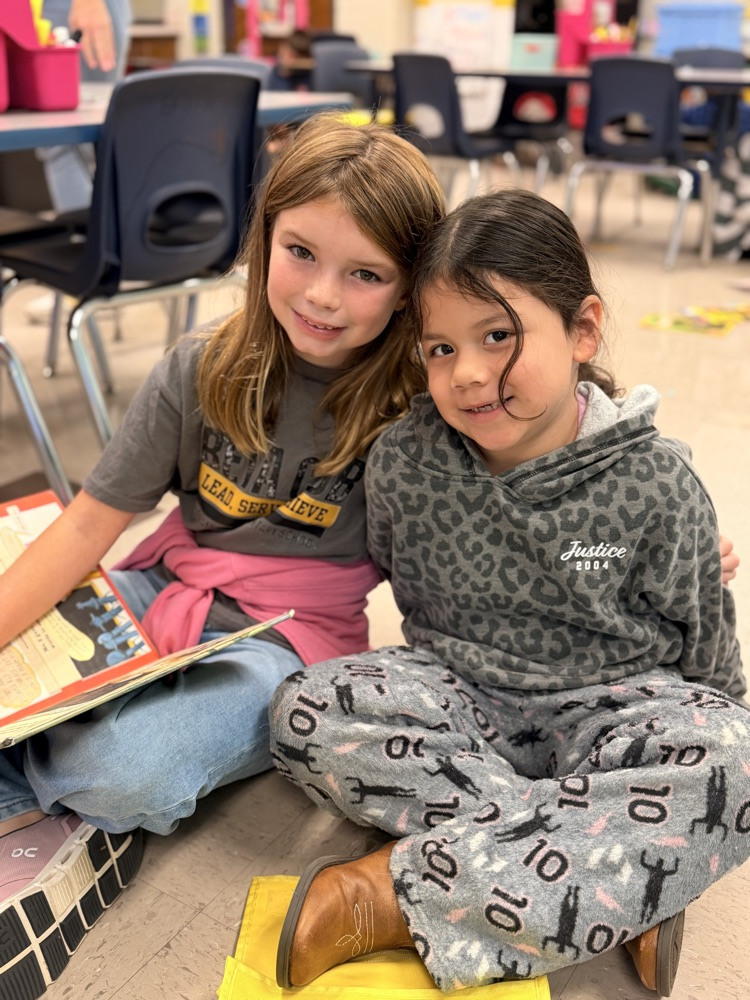 Two children sit on floor, one reads book, other looks at camera. Behind them, classroom with chairs, tables.