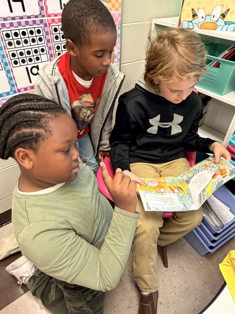 Three young children in a classroom, one reading a book while others listen attentively.