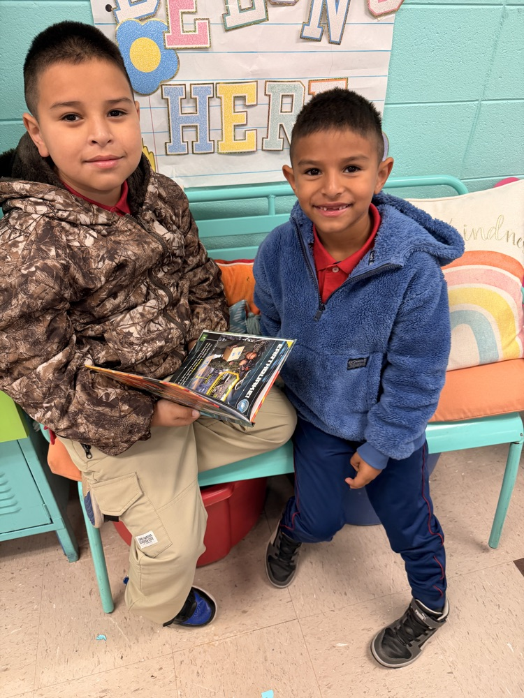 Two boys sit on a bench. The boy on the left holds a book. They both look at the camera.