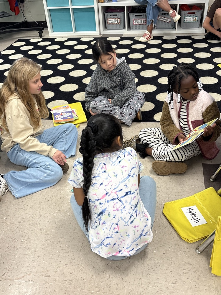 Four children sit cross-legged on a floor, surrounded by books, bags, and storage bins.