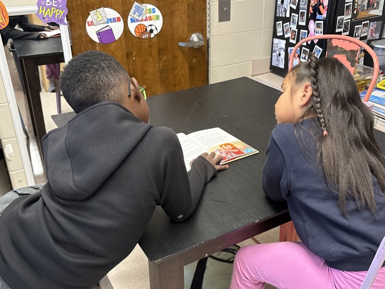 Two students seated at a table, one reading aloud from a book while the other listens attentively.