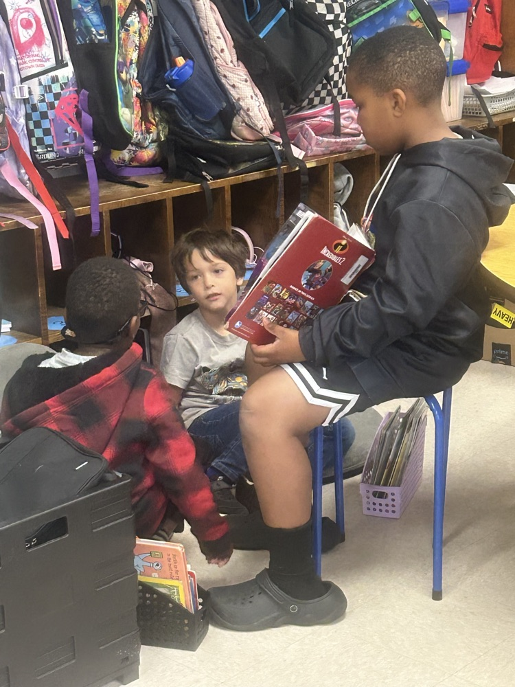 Students sitting in a classroom, one holding a comic book, another reaching into a box.