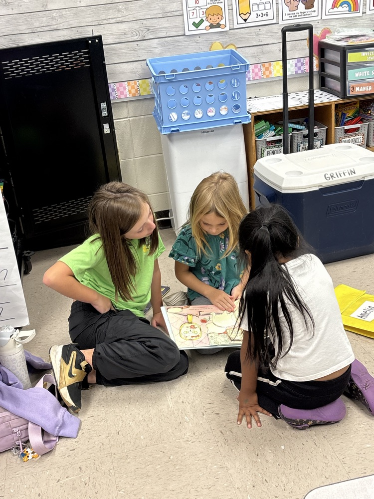 Three girls sitting on floor looking at picture in book; blue baskets and coolers in background.