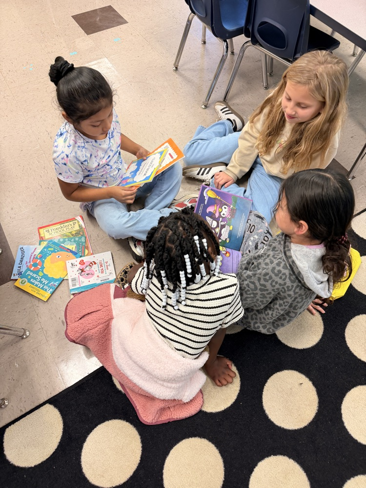 Four girls sit on a rug, reading books and smiling. A carpeted floor with round dots.