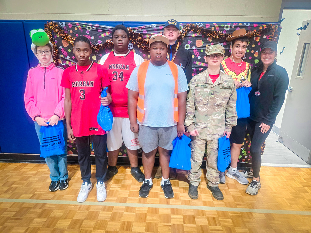 Several students and adults standing in a line in a school gym, smiling and holding bags.