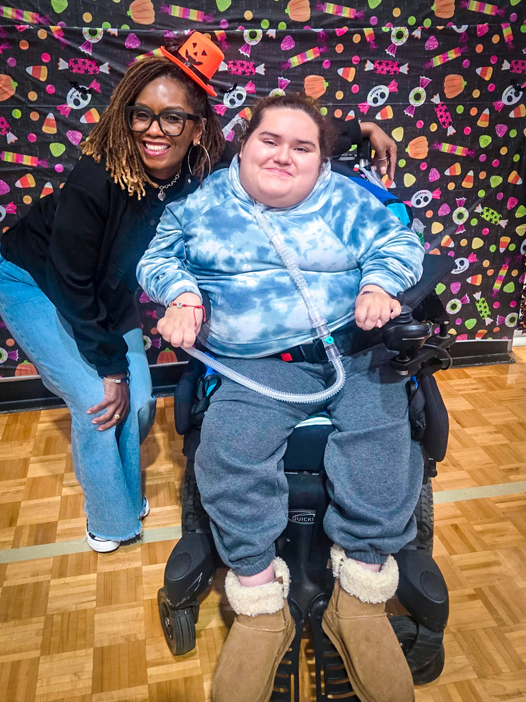 Two people stand behind a person in a wheelchair; the wheelchair has a tube connected to their nose.