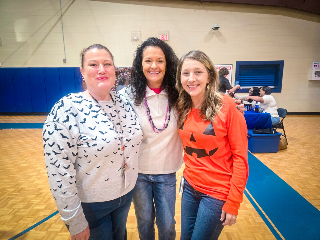 Three women wearing Halloween shirts in a gymnasium with a blue floor and a blue wall.