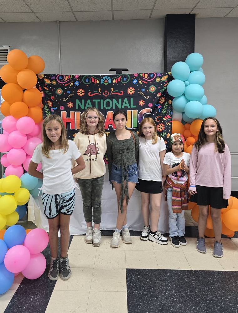 Picture with colorful balloons, and 6 female students standing in front of National Hispanic heritage banner.