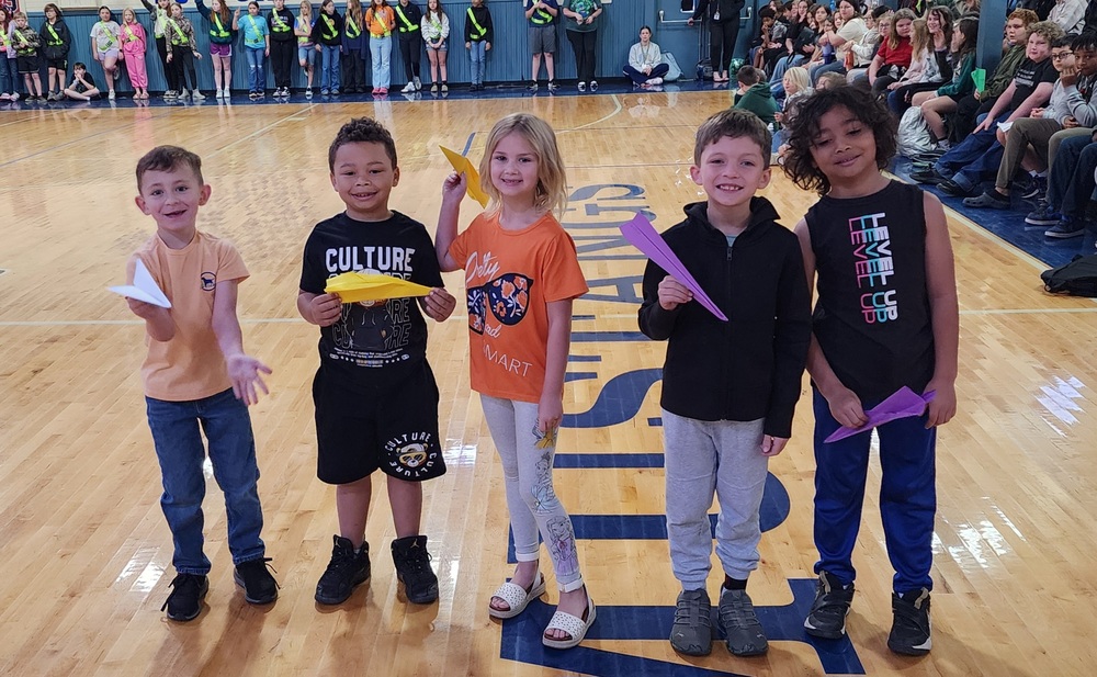 picture of 5 students, 4 boys and one girl holding paper airplanes in the gym