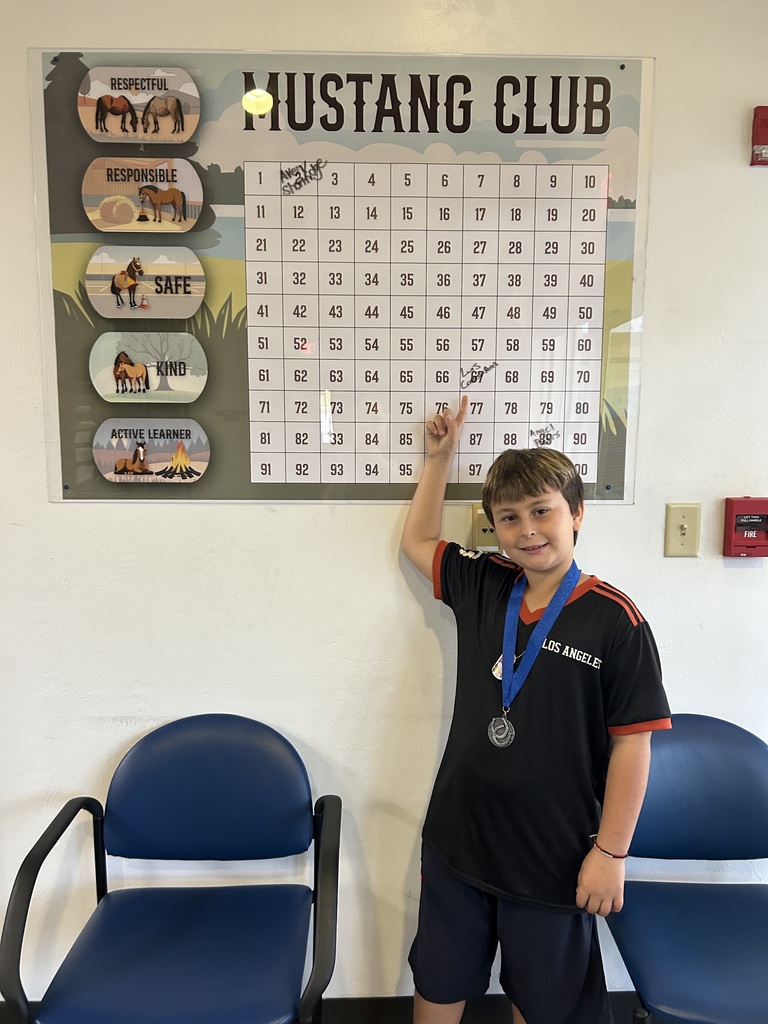 Picture of male student pointing at the Mustang Club board with his medal.