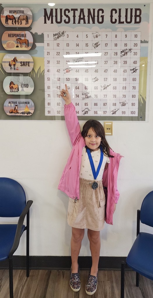 Female student wearing medal, pointing to the Mustang Club board.