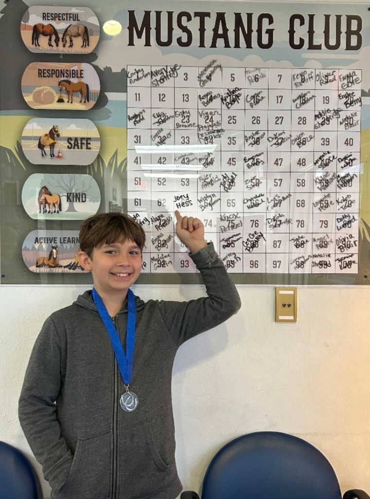 Picture of male student pointing at the Mustang Club board with his medal.