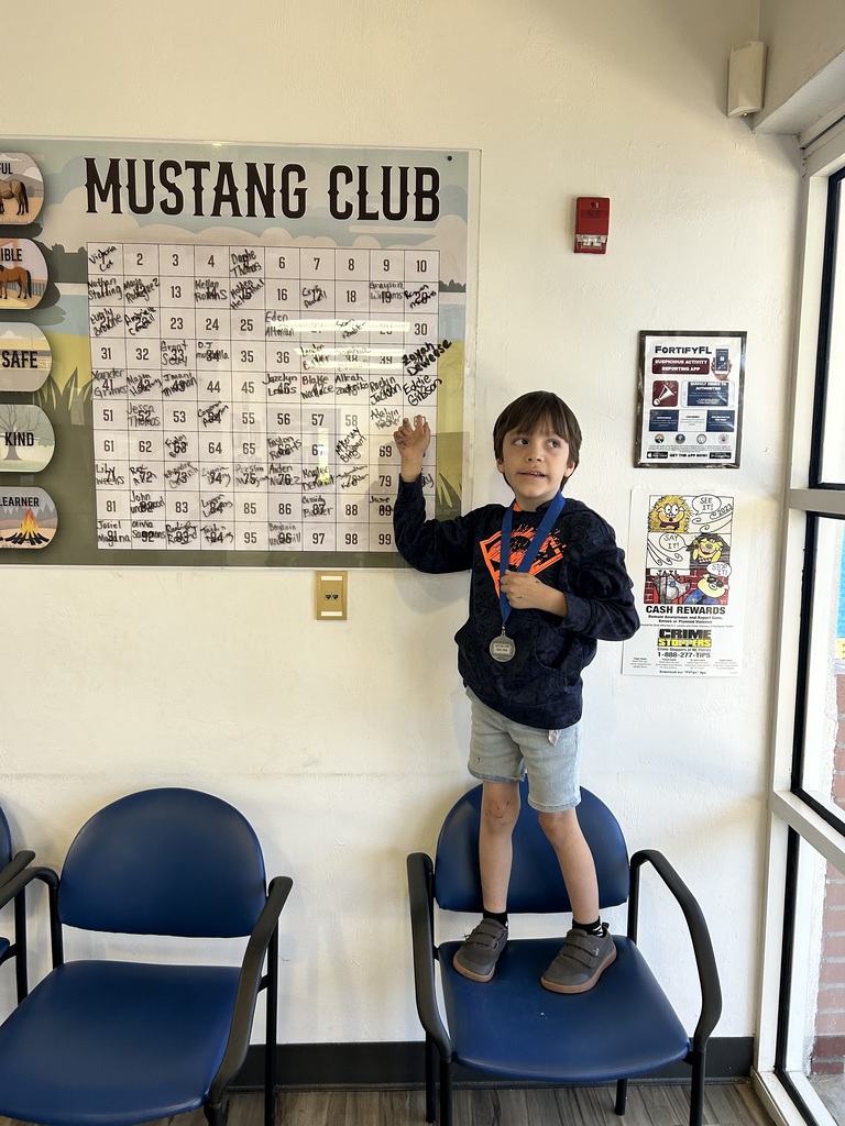 Picture of male student pointing at the Mustang Club board with his medal.