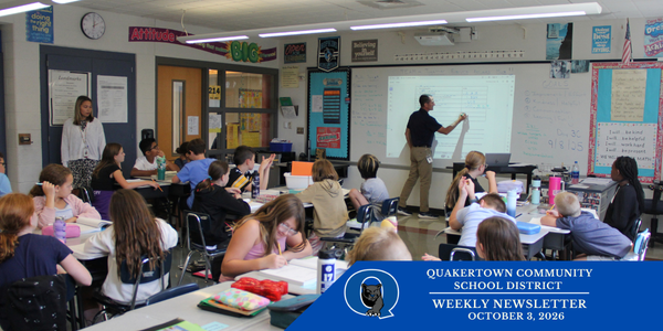 A photo of a classroom filled with students working hard at their desks and a teacher at the whiteboard instructing