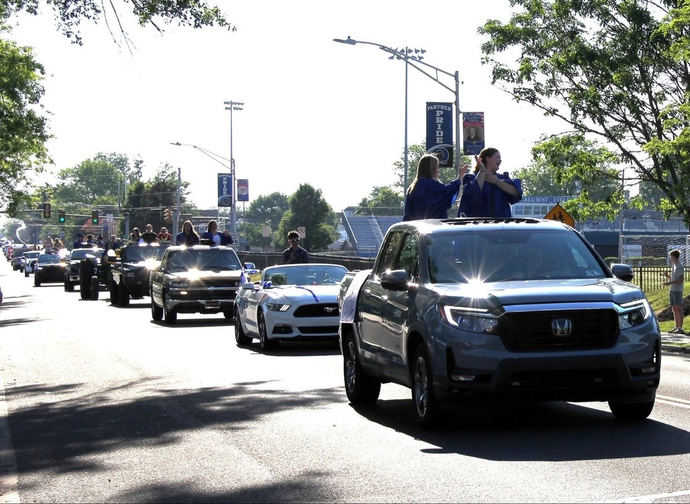 Image of decorated cars slowly driving dow the street with seniors waving from sun roofs and windows for the 2026 Parade of Graduates