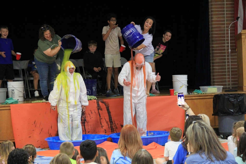 Two teachers, each standing in a blue baby pool, and wearing a white coverall get slimed from head to toe