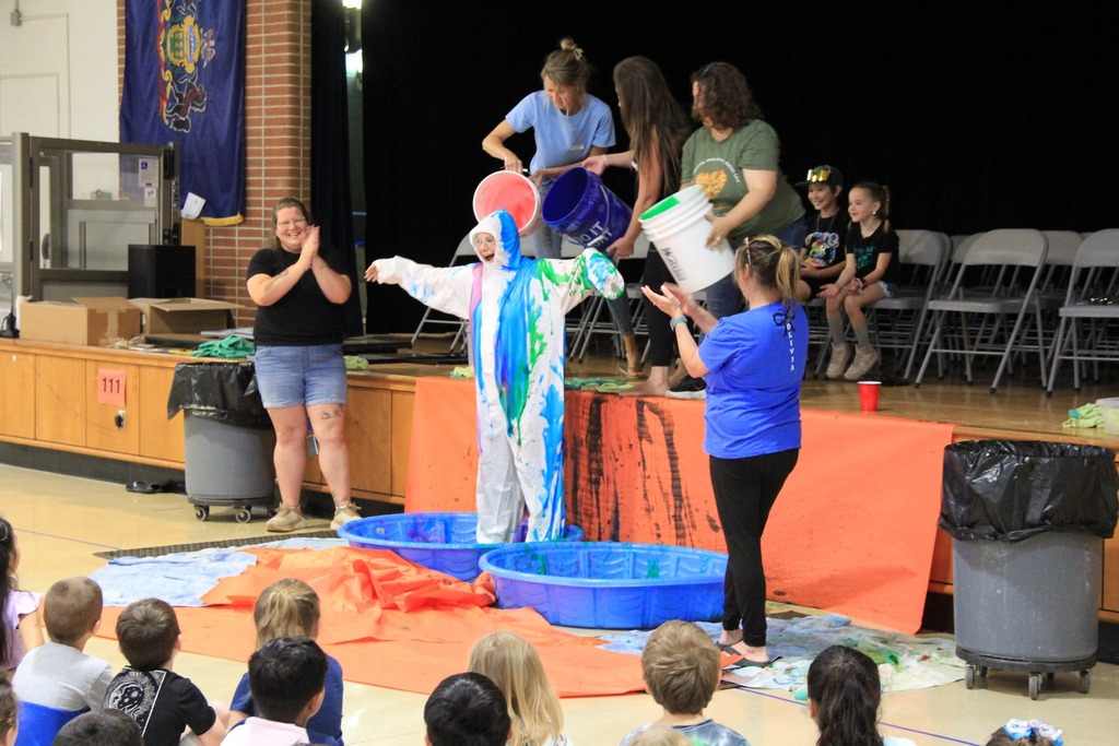 Dr. Z, standing in a baby pool and wearing a white coverall gets slimed from head to toe