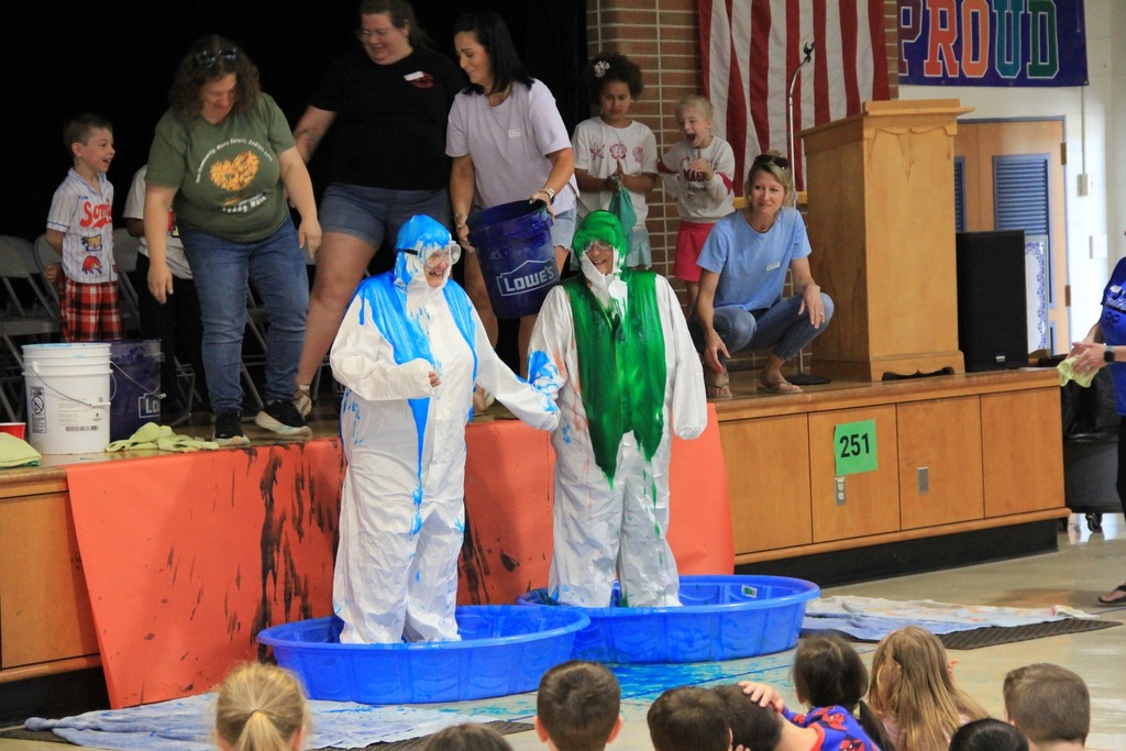 Two teachers holding hands, each standing in a blue baby pool, and wearing a white coverall get slimed from head to toe