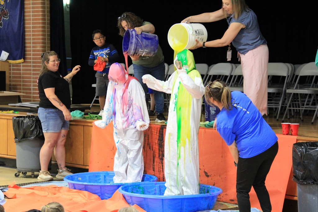 Two teachers, each standing in a blue baby pool, and wearing a white coverall get slimed from head to toe