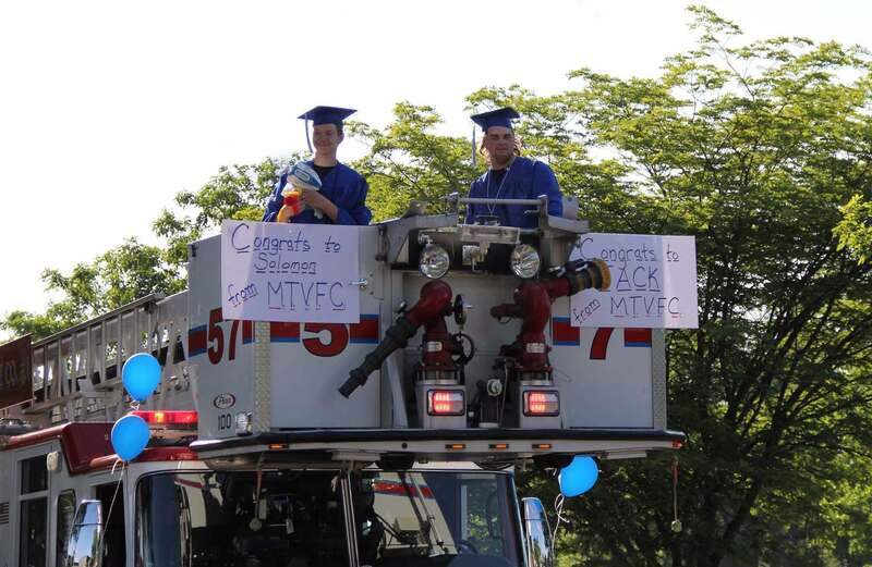 Students in caps and gowns waving to the crowd inside the ladder cady of a fire truck