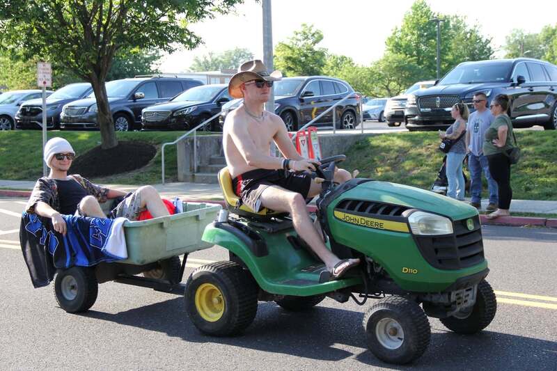 student rides a lawnmower, dragging a small trailer behind it with another student sitting