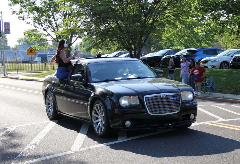Student sits on the outside of the window of a black car waving
